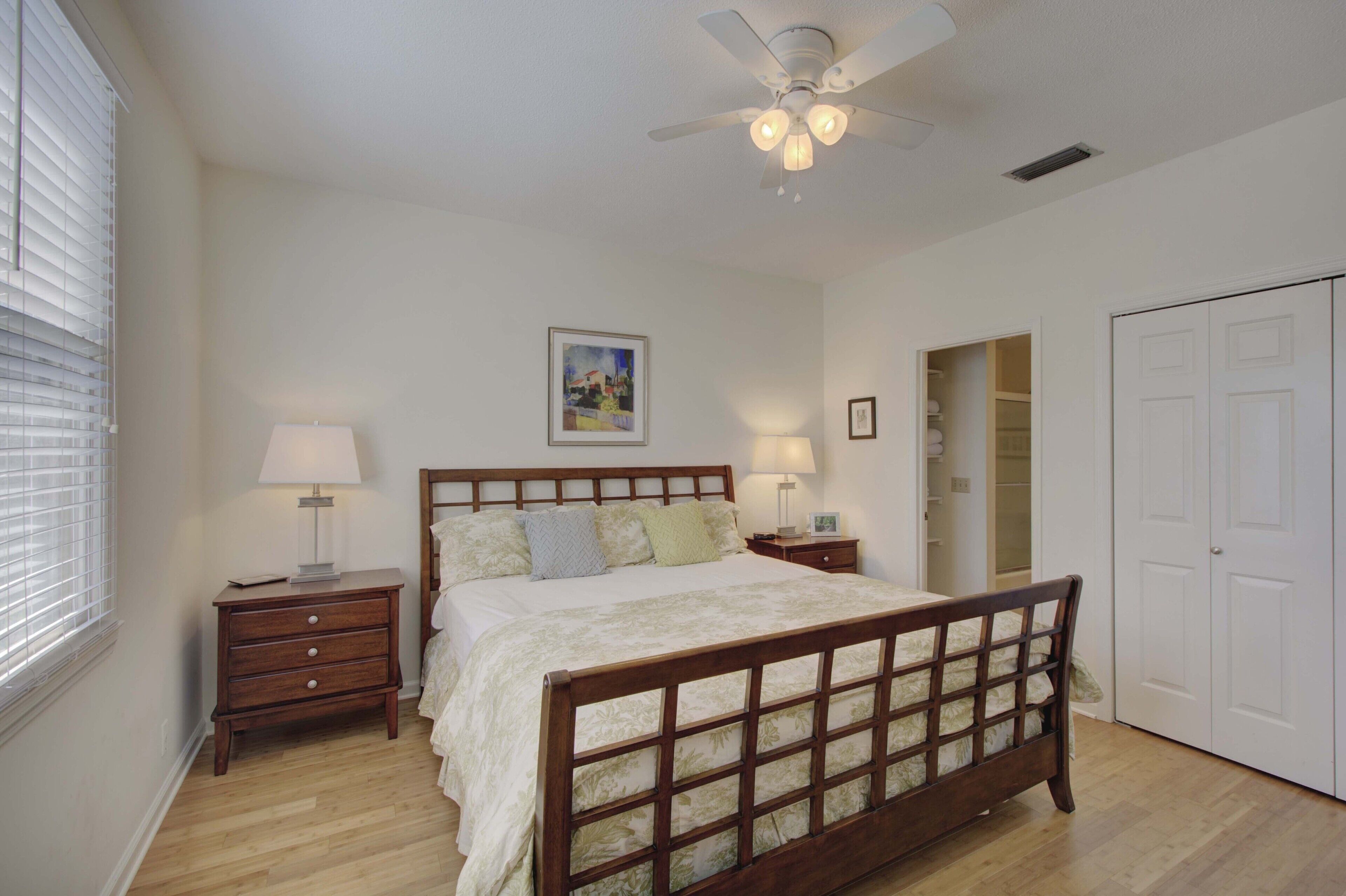 A bedroom with a wooden bed, two bedside tables with lamps, a ceiling fan, a closet, and a window with blinds.