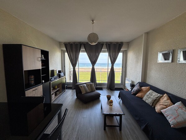 A living room with a sofa, coffee table, and TV stand. There is a view of the beach through large windows.