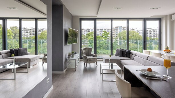 Een moderne woonkamer met een groot raam, een grijze bank, een salontafel en een eethoek met een glazen tafel.