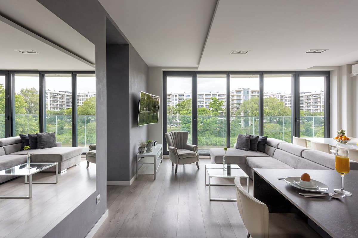 Een moderne woonkamer met een groot raam, een grijze bank, een salontafel en een eethoek met een glazen tafel.