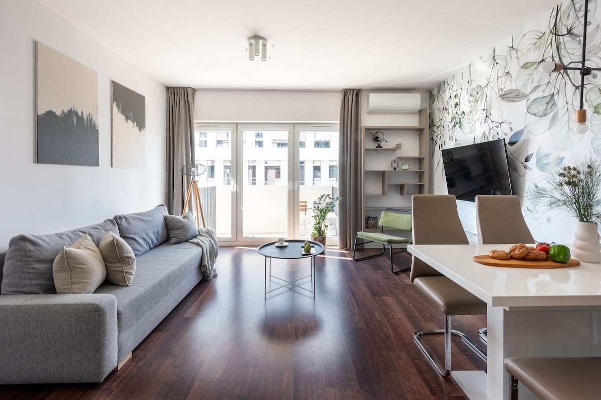 Een moderne woonkamer met een grijze bank, een ronde salontafel en een eethoek met een witte tafel en stoelen.