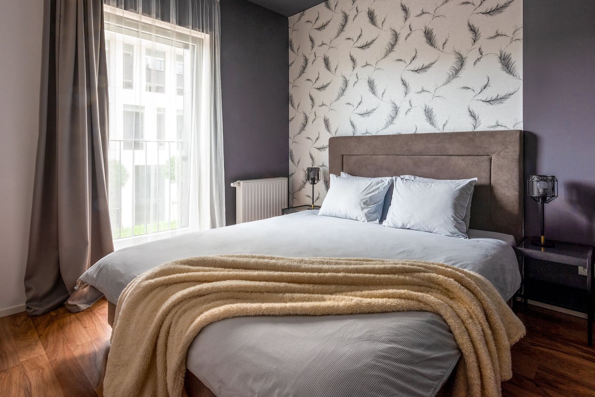 Een slaapkamer met een groot bed, een raam met gordijnen, een radiator en een muur met behang met een veermotief.