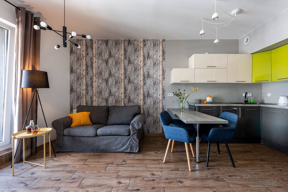 Een moderne woonkamer met een grijze bank, een bijzettafeltje met een lamp en een eethoek met blauwe stoelen.
