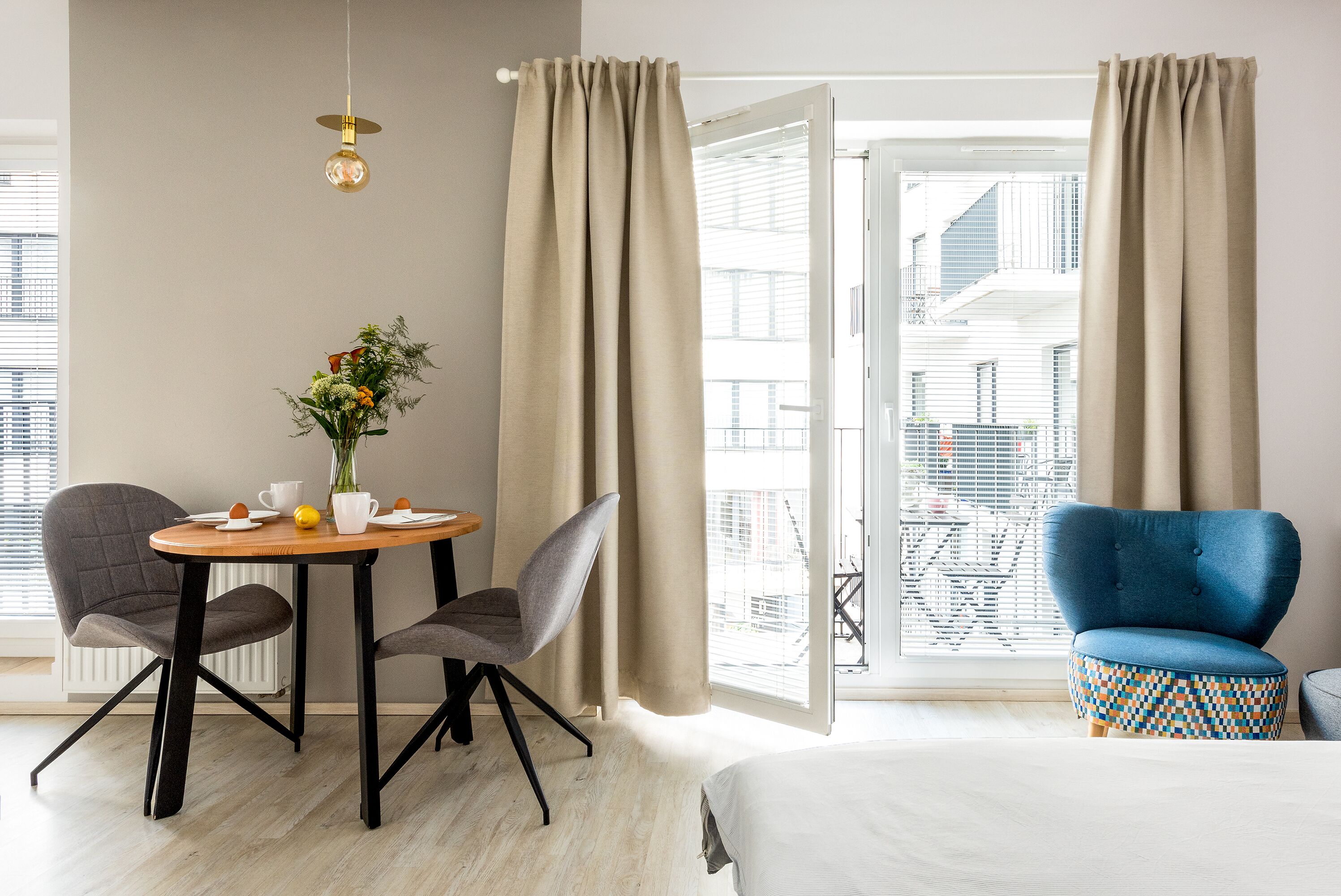 Ein Hotelzimmer mit einem Bett, zwei Stühlen, einem kleinen Tisch mit Blumenvase und Blick auf die Stadt durch große Fenster.