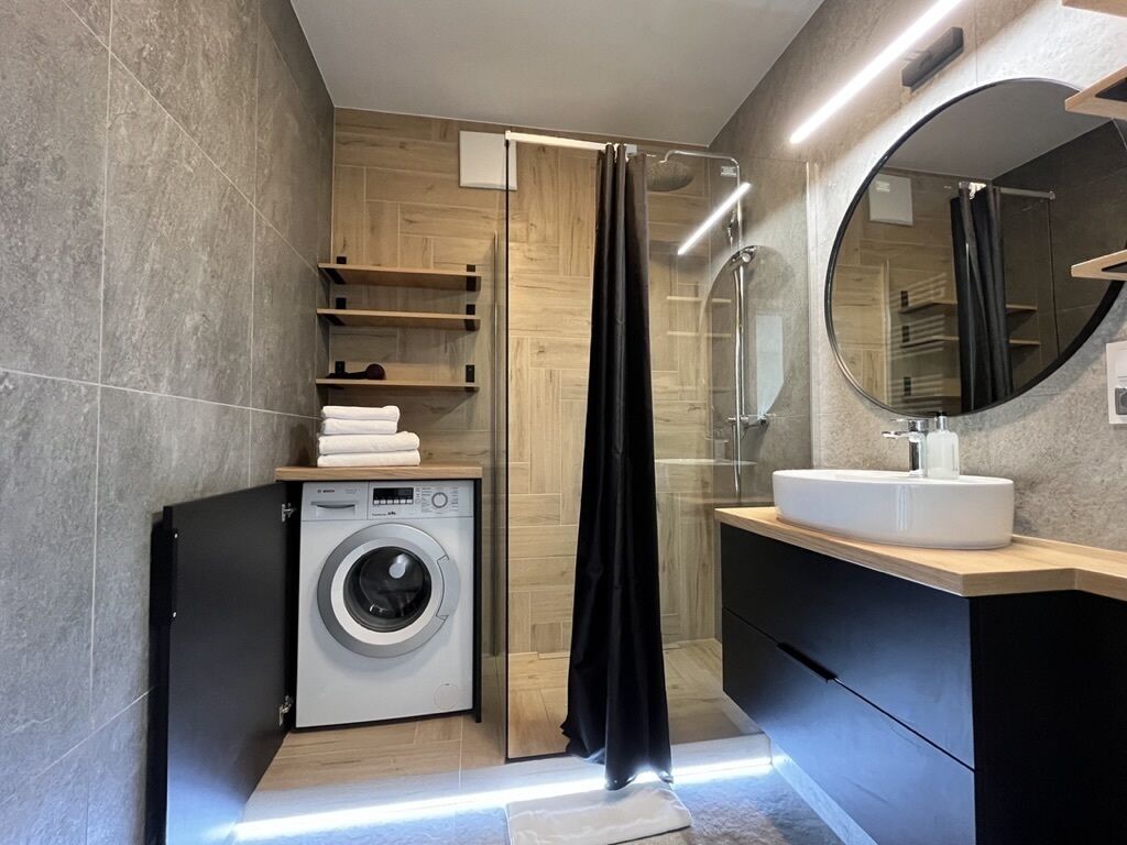 A modern bathroom with a washing machine, a shower with a glass door, and a round mirror above the sink.