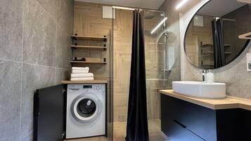 A modern bathroom with a washing machine, a shower with a glass door, and a round mirror above the sink.
