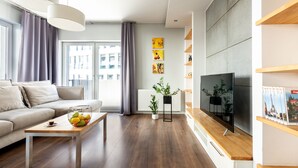 Een moderne woonkamer met een bank, salontafel en televisie. Er staan planten, een radiator en een raam met gordijnen.