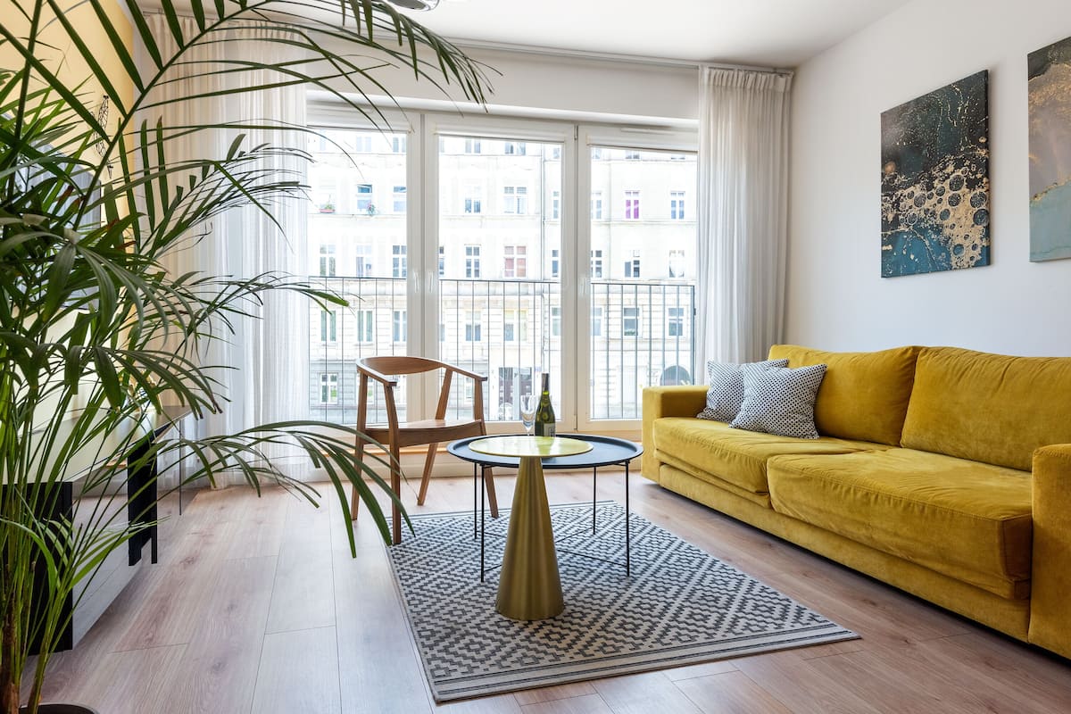 Een moderne woonkamer met een gele bank, een ronde salontafel en een groot raam met uitzicht op gebouwen.