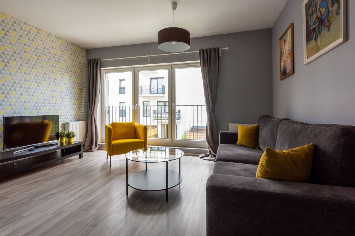 Een moderne woonkamer met een bank, fauteuil en salontafel, een televisie en een raam met uitzicht op gebouwen.