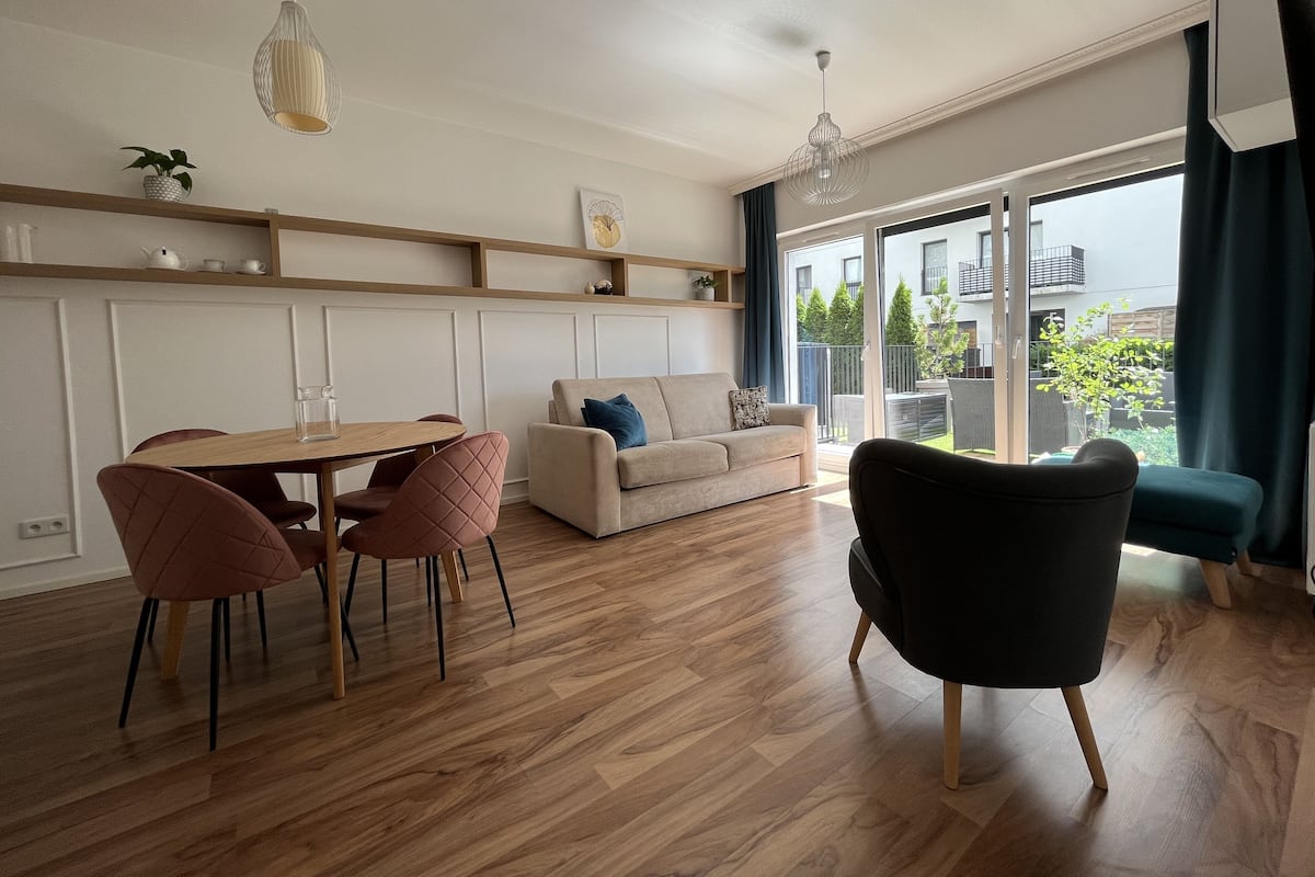 Een moderne woonkamer met een houten eettafel, twee banken en een zwarte fauteuil. Er zijn hanglampen, een salontafel en een uitzicht op het buitenlandschap door grote ramen.