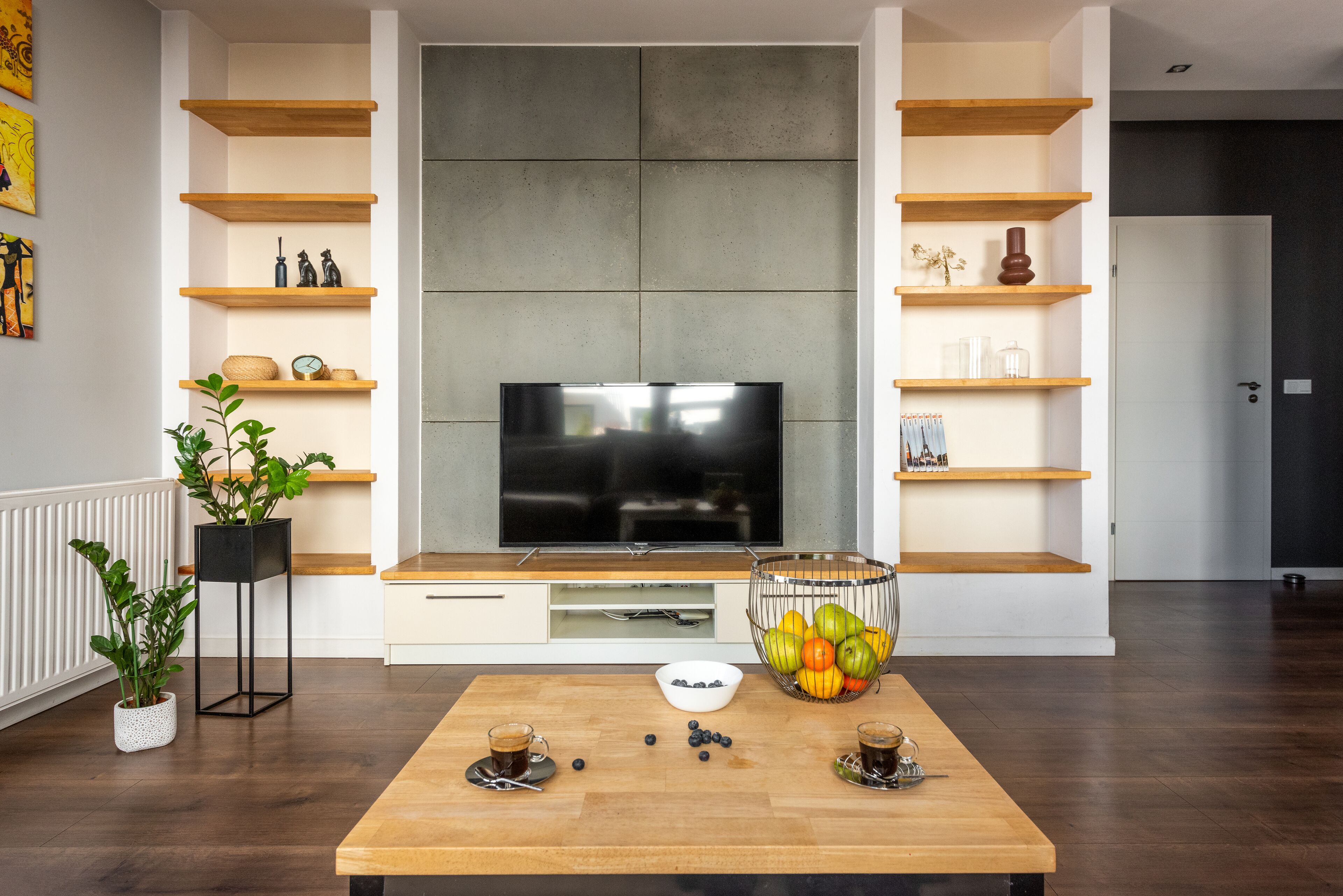 Una sala de estar moderna con un televisor de pantalla plana, estantes de madera, una mesa de centro con frutas y tazas, y una planta en maceta.
