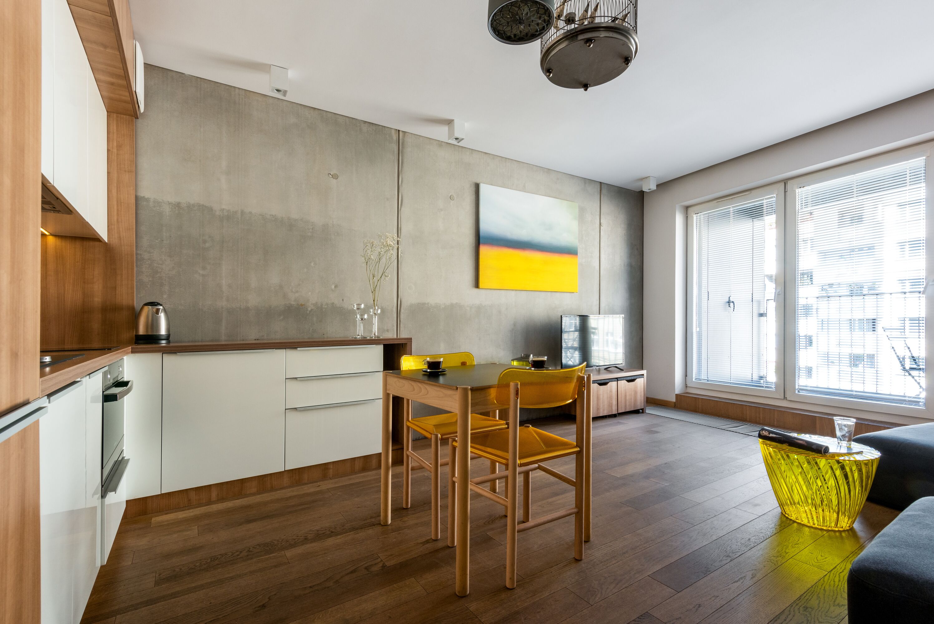 A modern kitchen with wooden cabinets, a dining table, and chairs. There is a yellow chair, a glass vase, and a yellow side table.