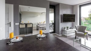 Een moderne woonkamer met een donkere vloer, een glazen salontafel, een grijze fauteuil en een eethoek met een glazen tafel en stoelen.