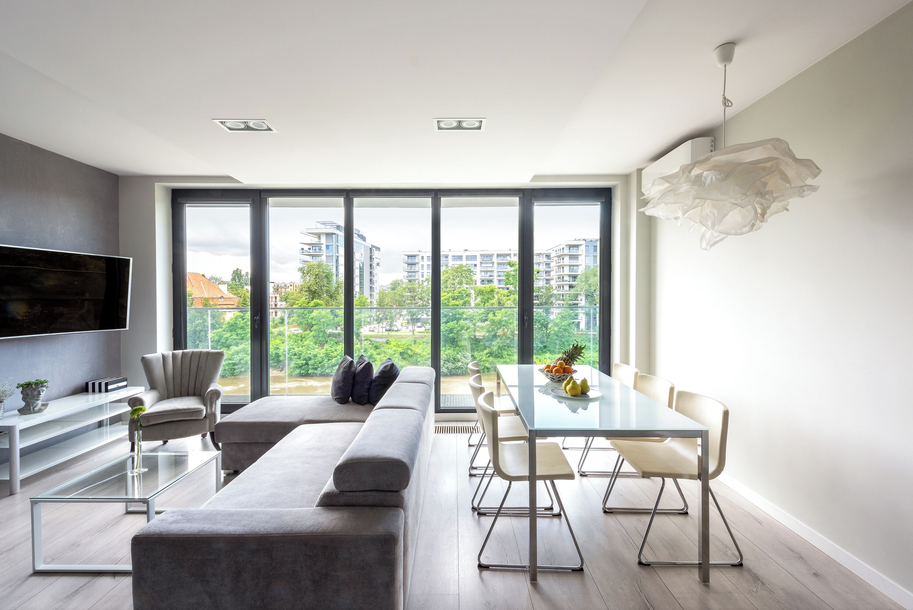 A modern living room with a large TV, comfortable seating, a dining area, and a view of buildings and greenery.