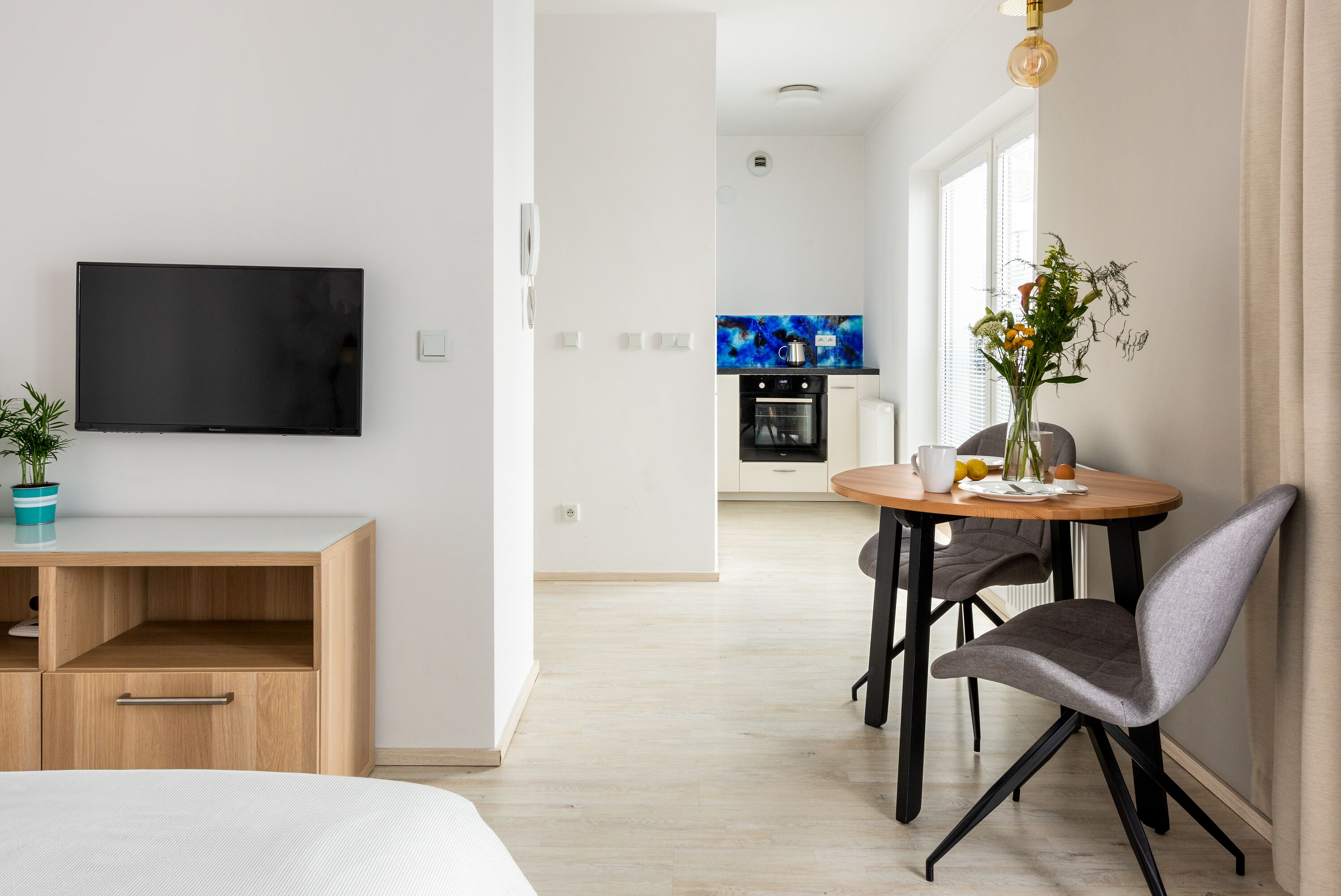 A modern living room with a flat-screen TV, a wooden desk, a dining area with a table and chairs, and a kitchen in the background.