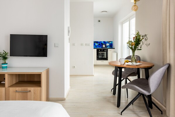 Una sala moderna con una televisión de pantalla plana, un escritorio de madera, una zona de comedor con mesa y sillas, y una cocina al fondo.