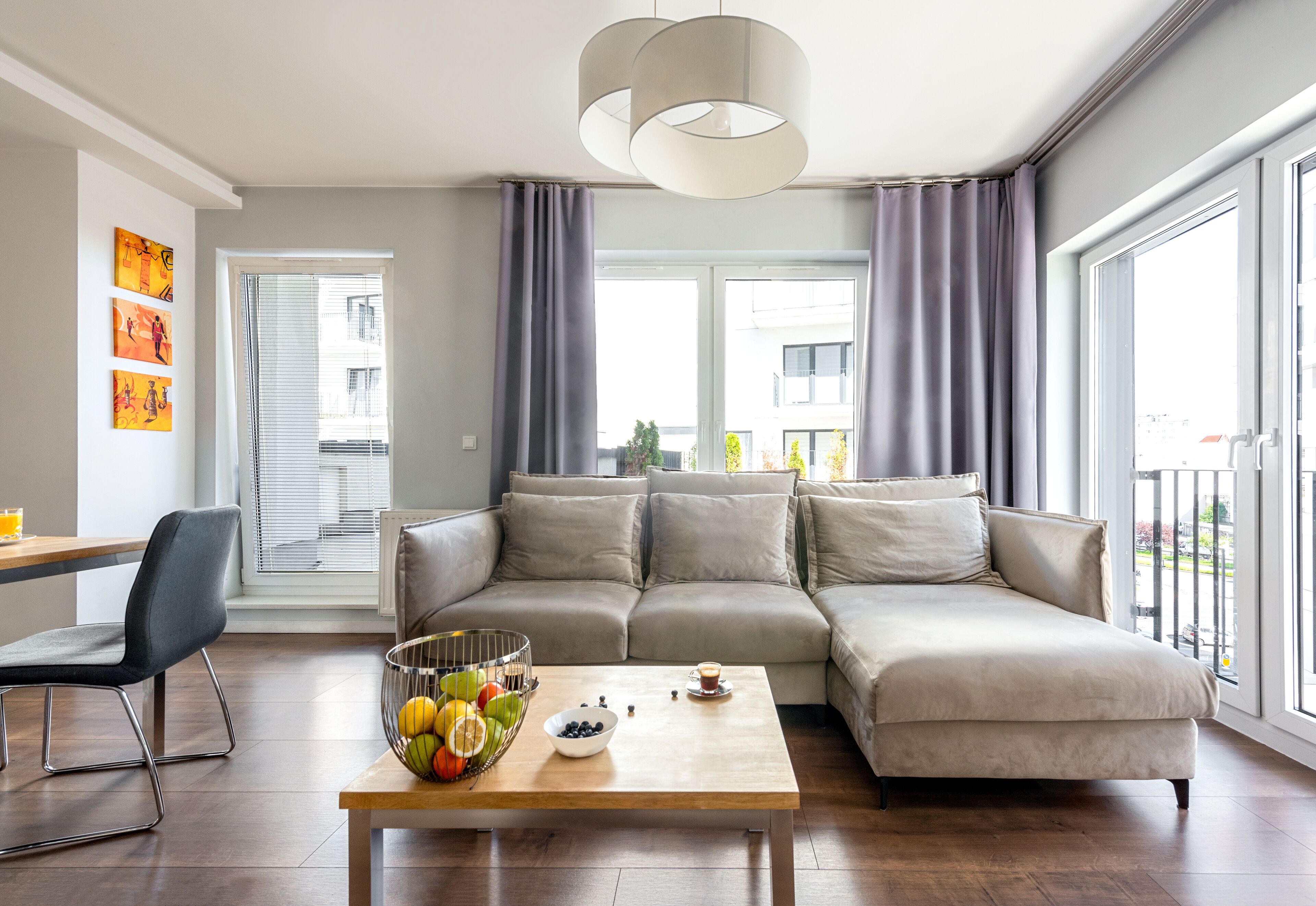 A modern living room with a sectional sofa, a dining area with a chair, a coffee table with fruits and a cup, and large windows with curtains.