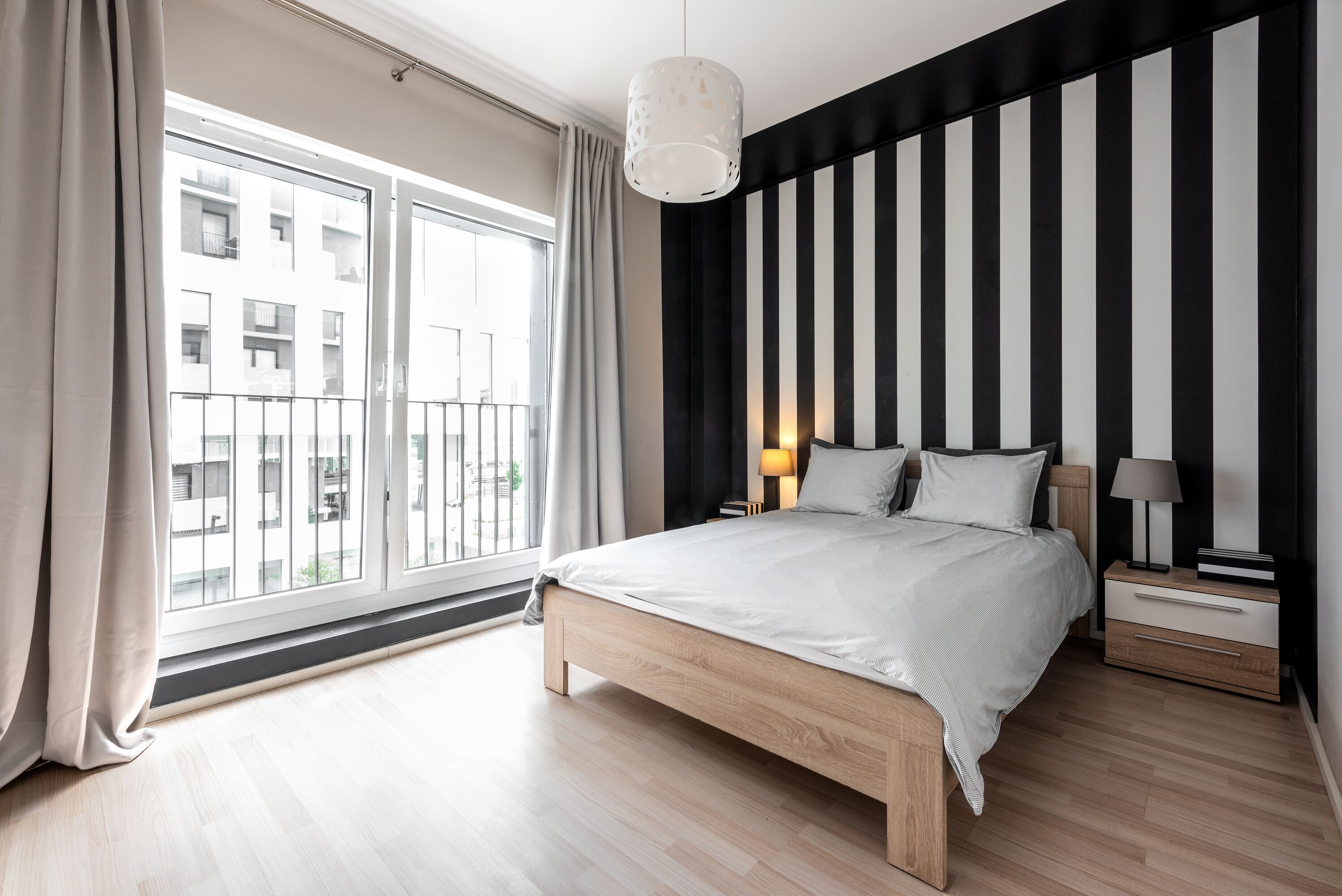 A modern bedroom with a large window, a bed, a nightstand, and a striped wall.