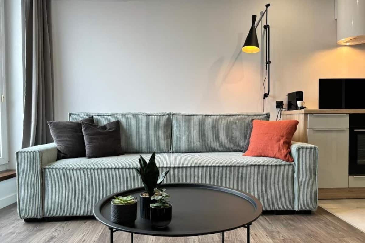 Een moderne woonkamer met een gestreept bankje, een ronde salontafel met planten, een zwarte vloerlamp en een keuken in de achtergrond.