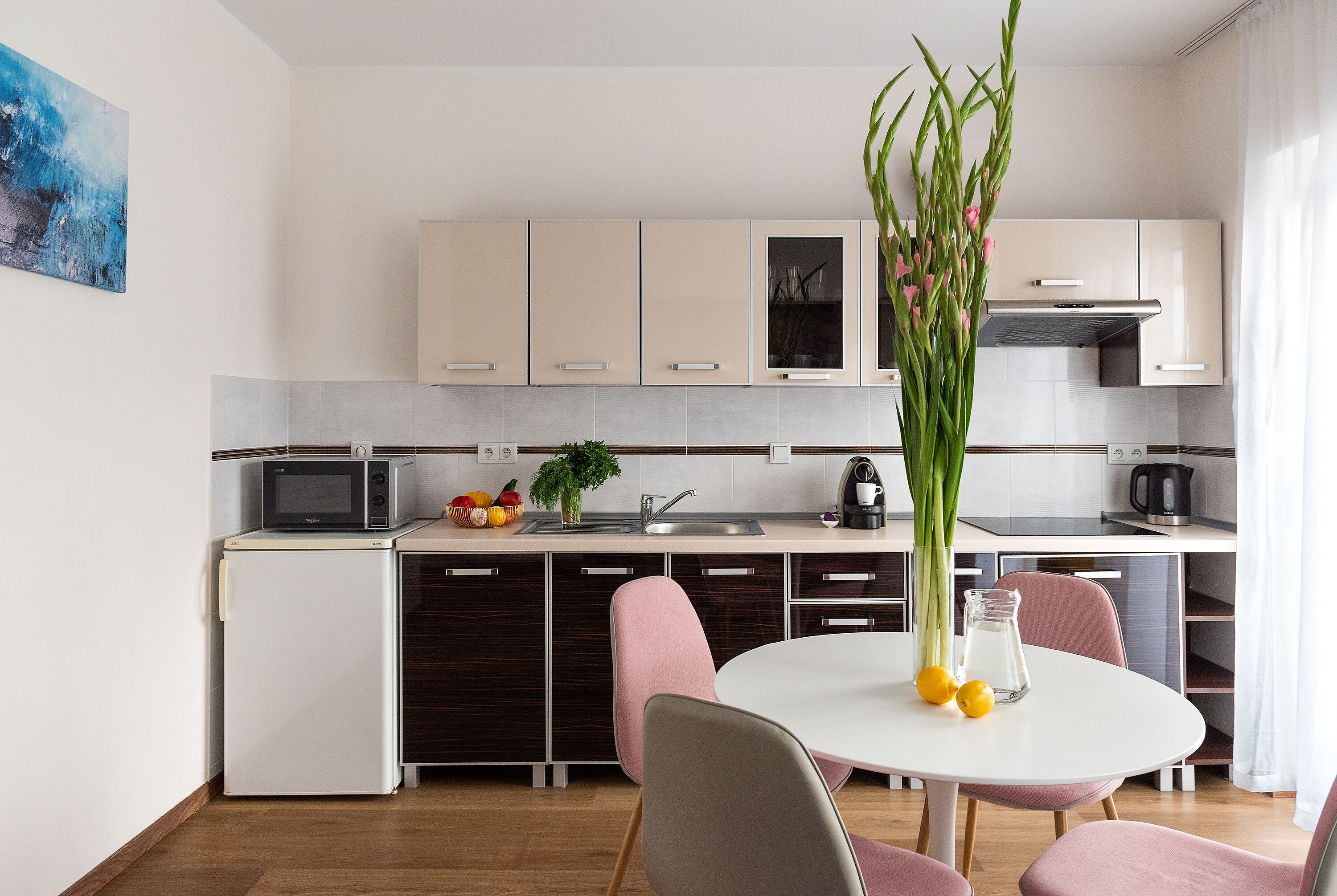 A modern kitchen with white cabinets, a microwave, a refrigerator, and a dining area with pink chairs and a white table.