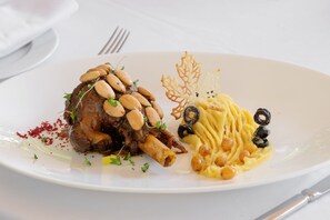 A white plate with a lamb shank, almonds, and a decorative leaf. - Pavillon Terra ababila (Marrakech)
