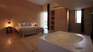 A bedroom with a large bed, a sitting area with a chair, a bathtub, and a small table with a lamp. - Pavillon Terra ababila (Marrakech)