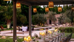 An outdoor dining area with tables set for a meal, surrounded by greenery and palm trees. - Pavillon Terra ababila (Marrakech)