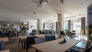 A spacious dining area with multiple tables and chairs, a kitchen in the background, and large windows offering a view of the outdoors. - Heuvelrijk Berg en Dal (Groesbeek)