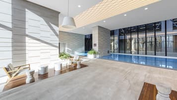 Área moderna de piscina interna com piso de madeira, assentos e vista para o exterior.