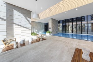 Área moderna de piscina interna com piso de madeira, assentos e vista para o exterior.