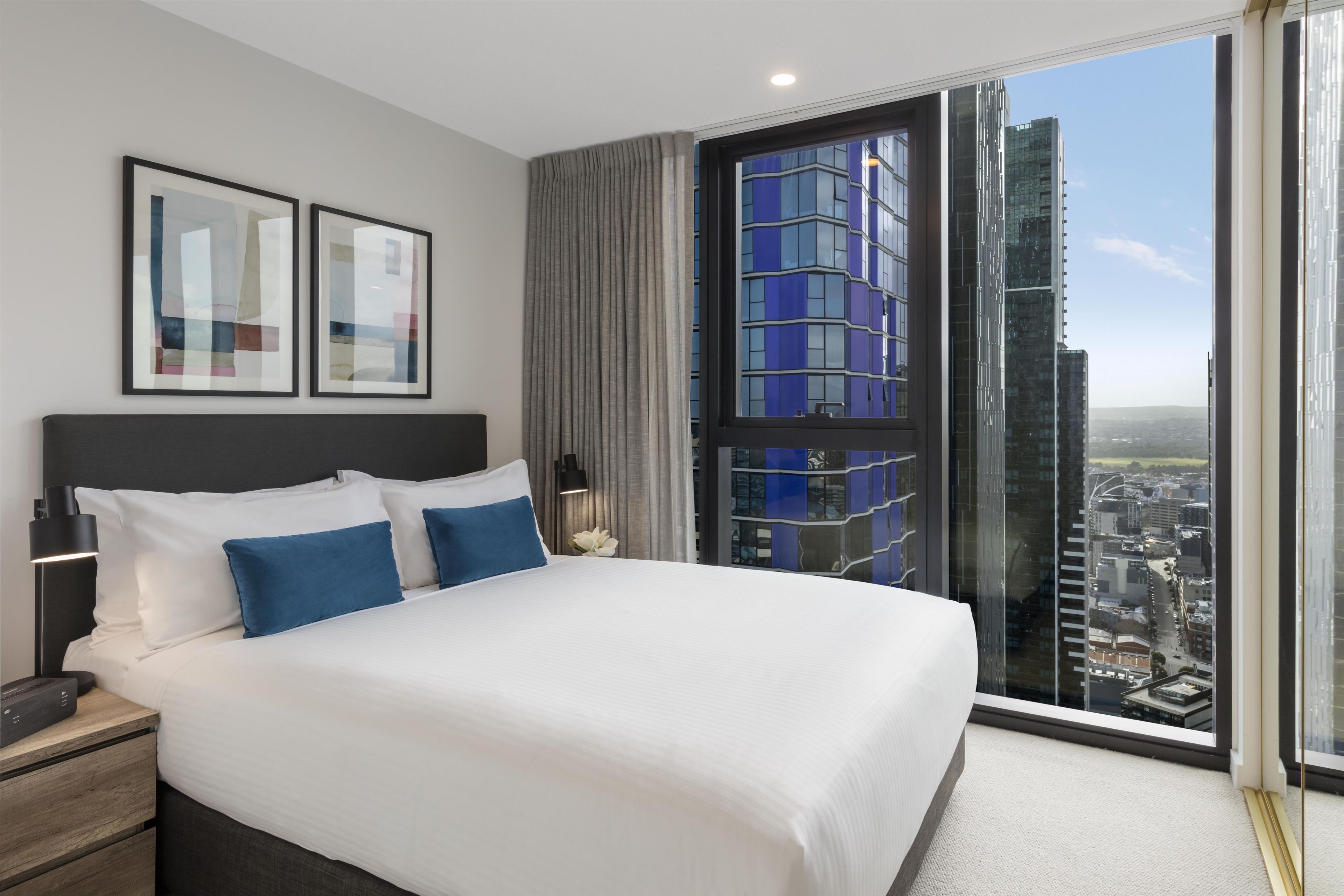 A modern hotel room with a large bed, a nightstand, a lamp, and a view of a cityscape through a large window.