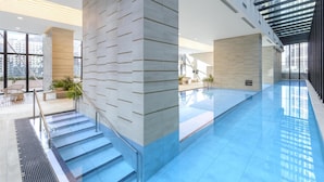 Piscina interior moderna com paredes de vidro transparente, chão em azulejo e escada com guarda-corpos de metal.