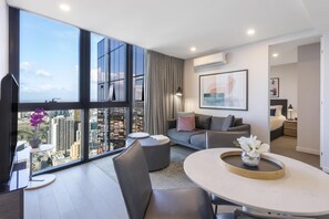 A modern living room with a large window, a round table, a sofa, and a bed in the adjacent room.