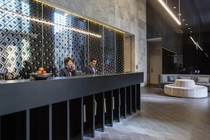 A reception area with a modern design, featuring a patterned wall, a reception desk, and a seating area with a sofa and ottoman. - Avani Melbourne Central Residences (Melbourne)