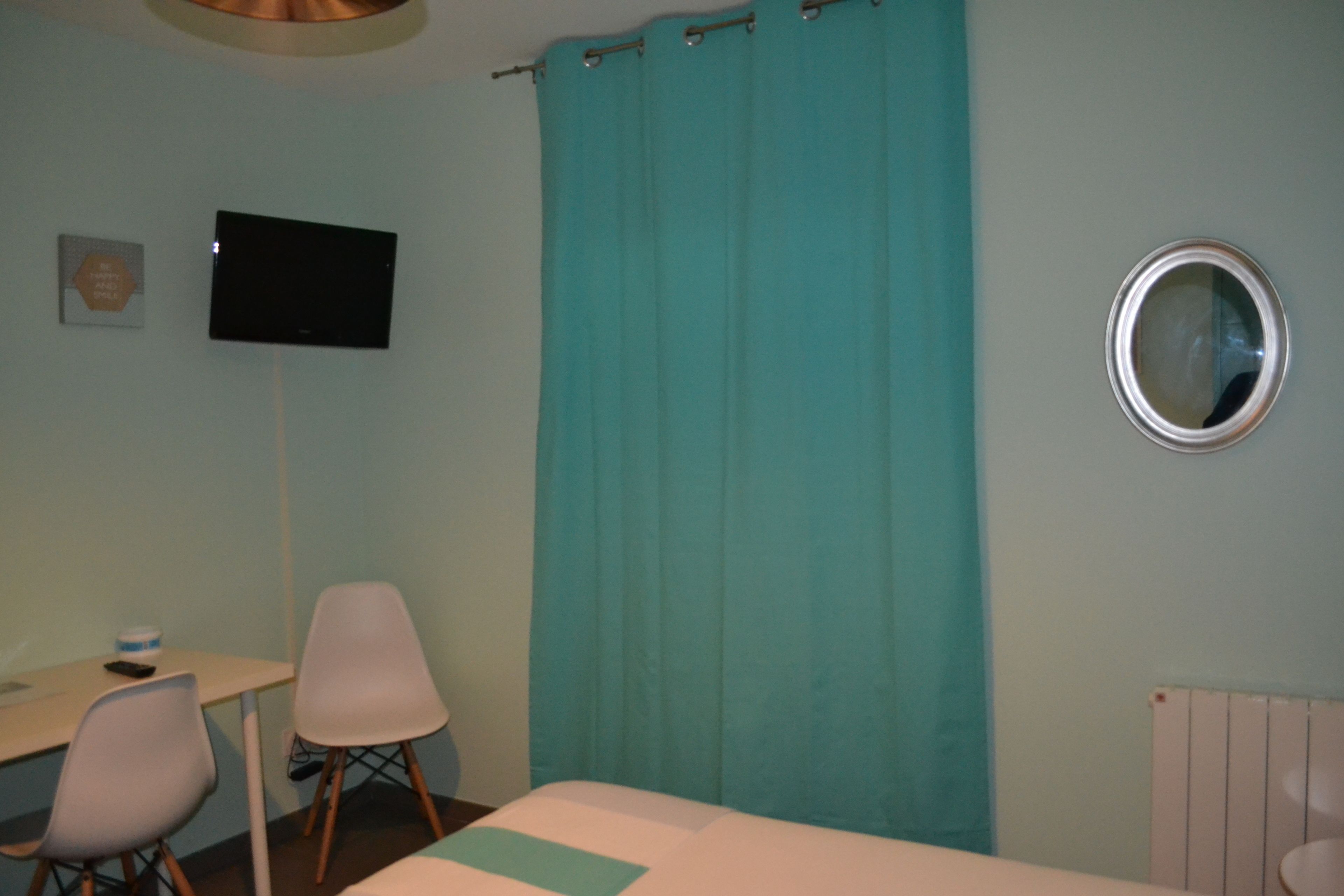 A room with a bed, a desk, two chairs, a television, a teal curtain, and a round mirror on the wall.