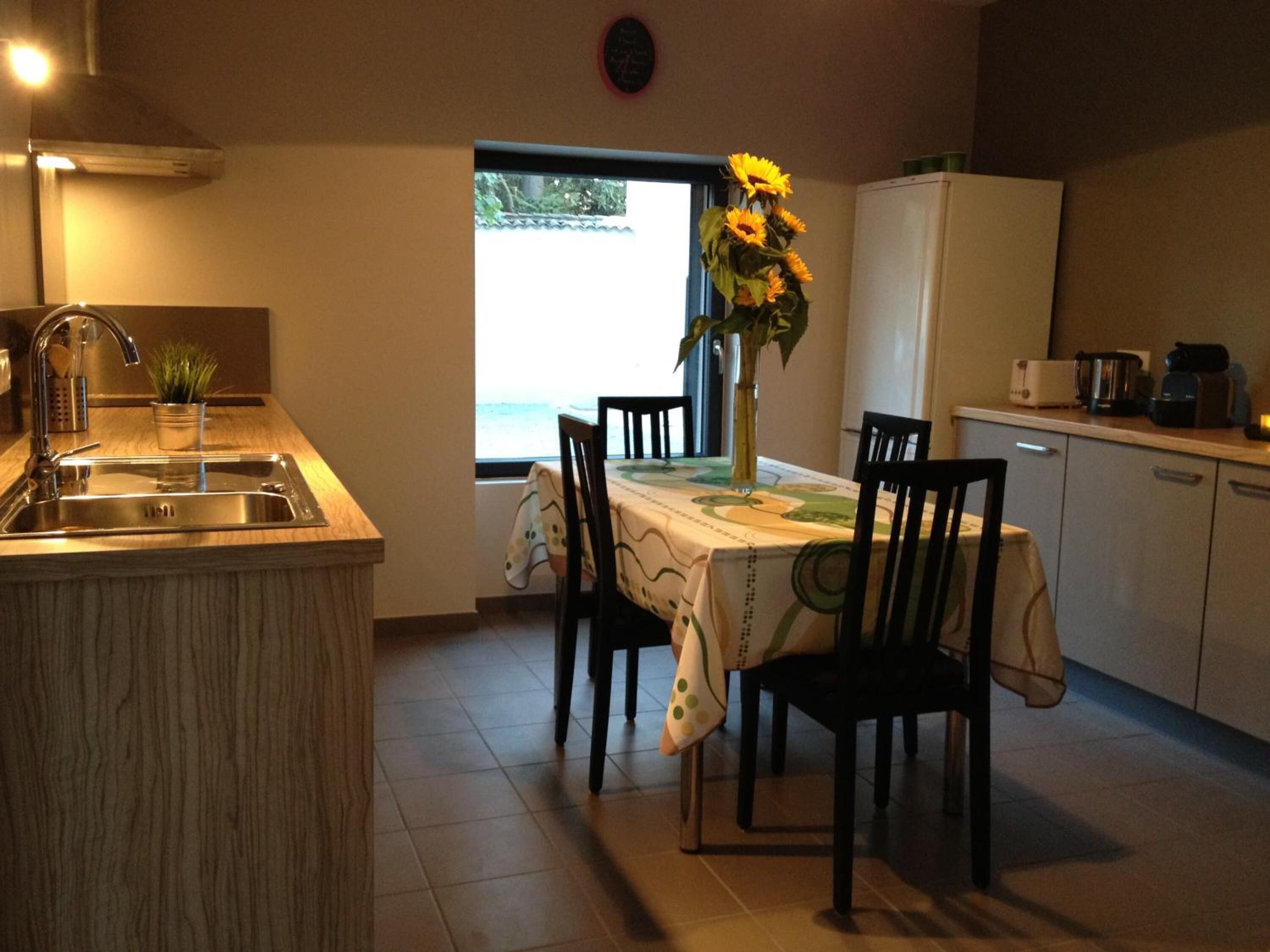 A compact kitchen with a sink, a dining table with chairs, a vase with sunflowers, and a view of the outdoors through a window.