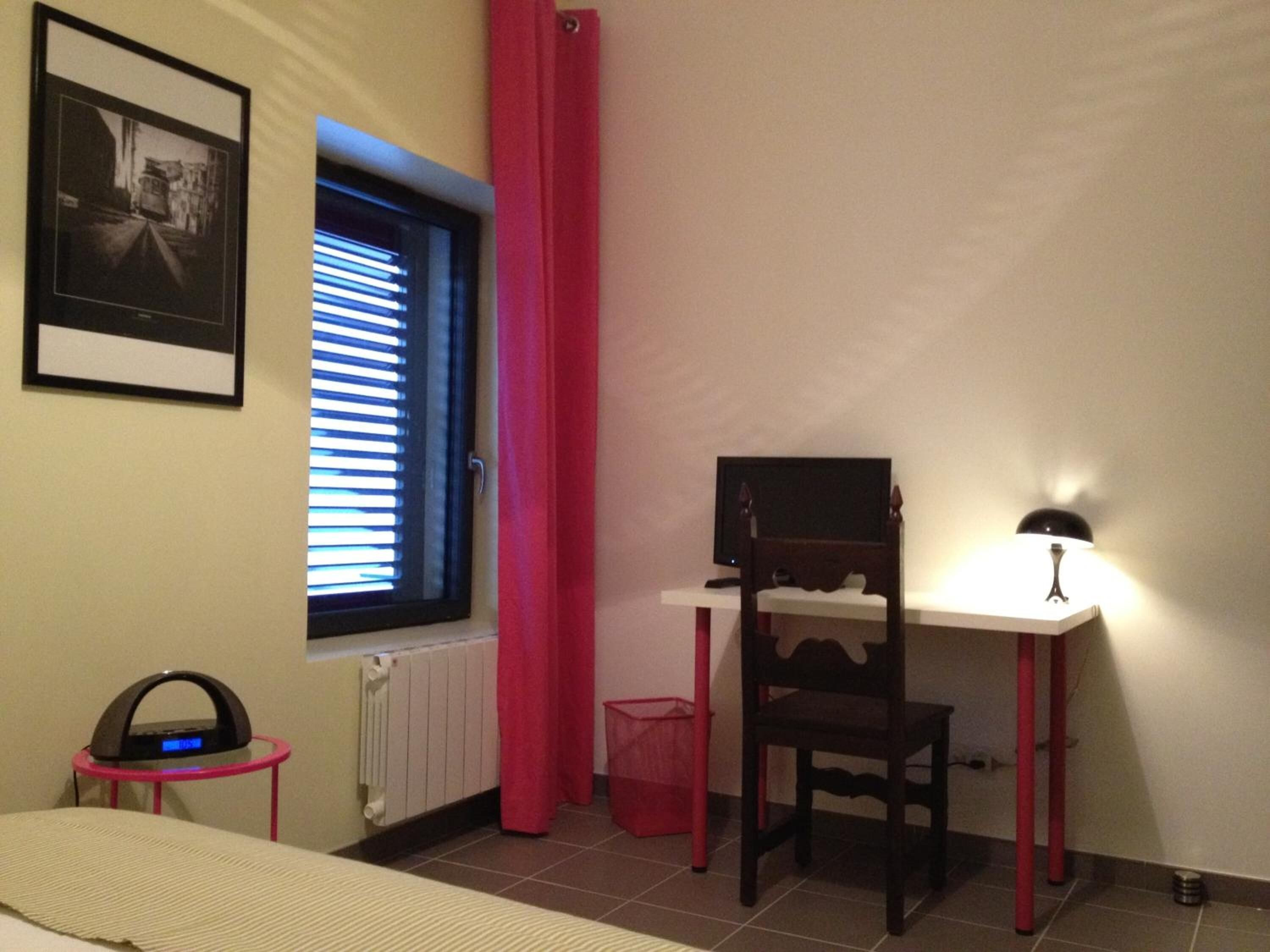 A small, well-lit room with a bed, a desk with a chair, a radiator, a window with blinds, a framed picture on the wall, and a red trash bin.