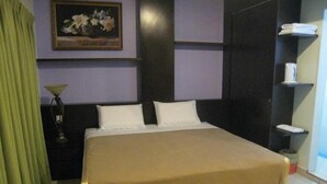 Uma cama bem arrumada com cobertura bege, um criado-mudo com abajur e um quadro na parede.