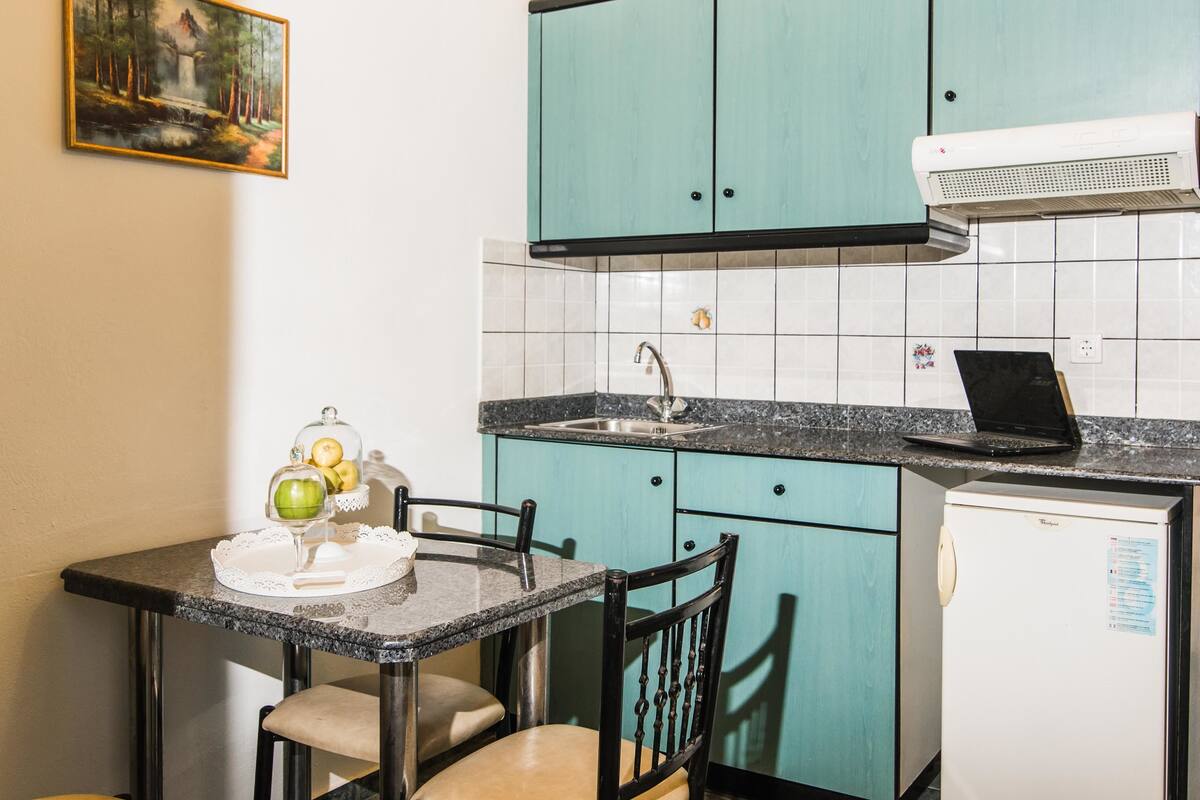 Een kleine keuken met blauwgroene kastjes, een spoelbak, een fornuis en een koelkast. Op het aanrecht ligt een laptop.