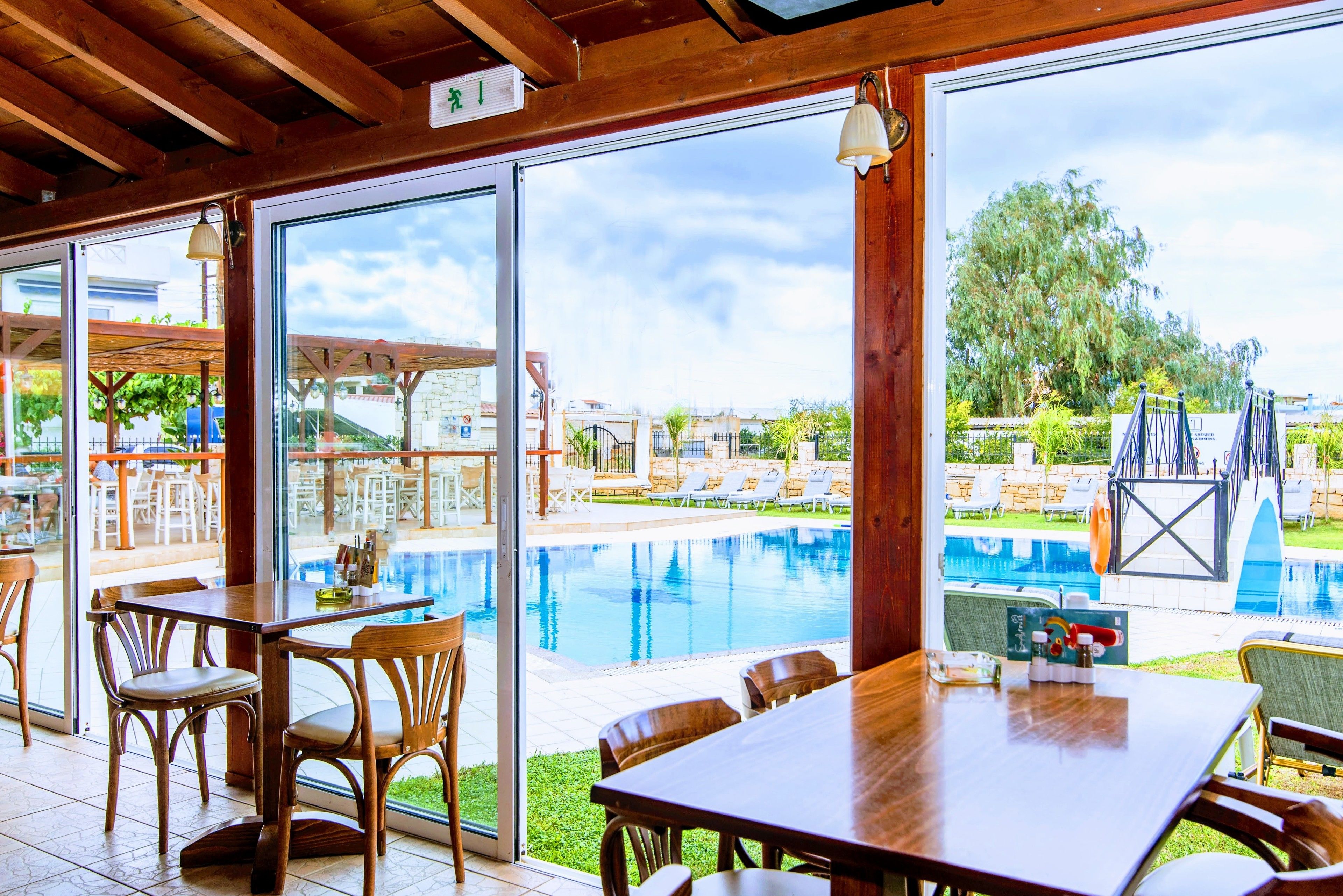A poolside area with wooden tables and chairs, a clear view of the pool, and a covered seating area with umbrellas.