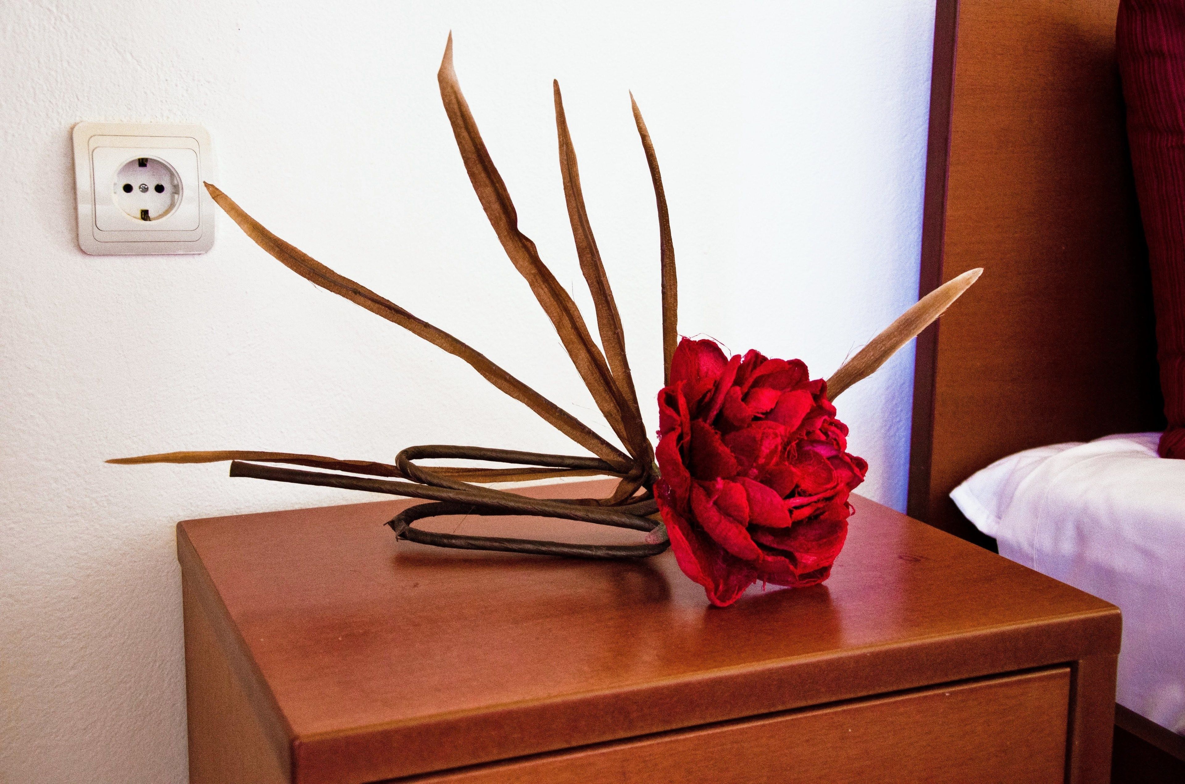 A wooden bedside table with a red flower arrangement and a power outlet on the wall.