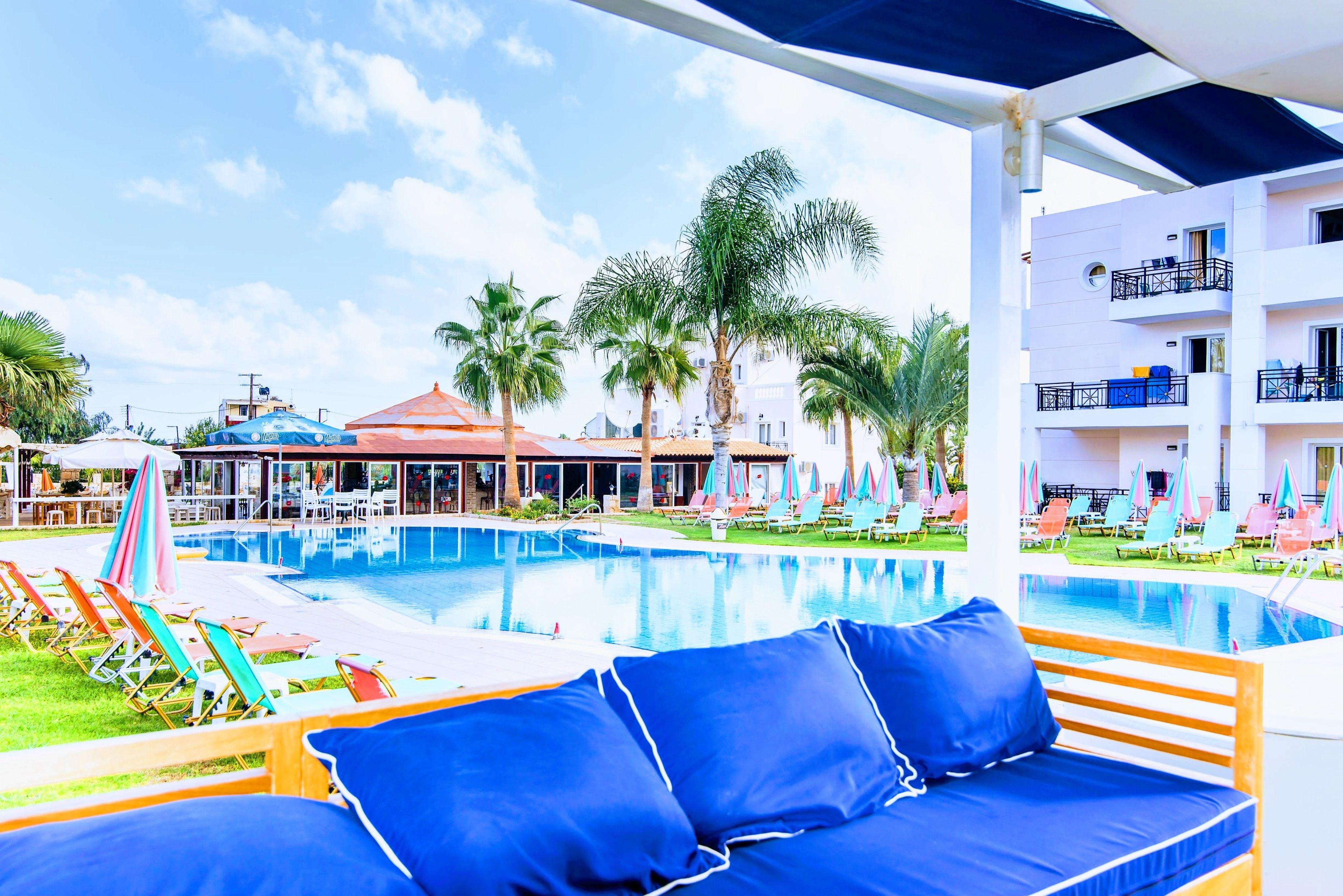 A pool area with lounge chairs and umbrellas, a covered seating area with blue cushions, and a building with balconies.