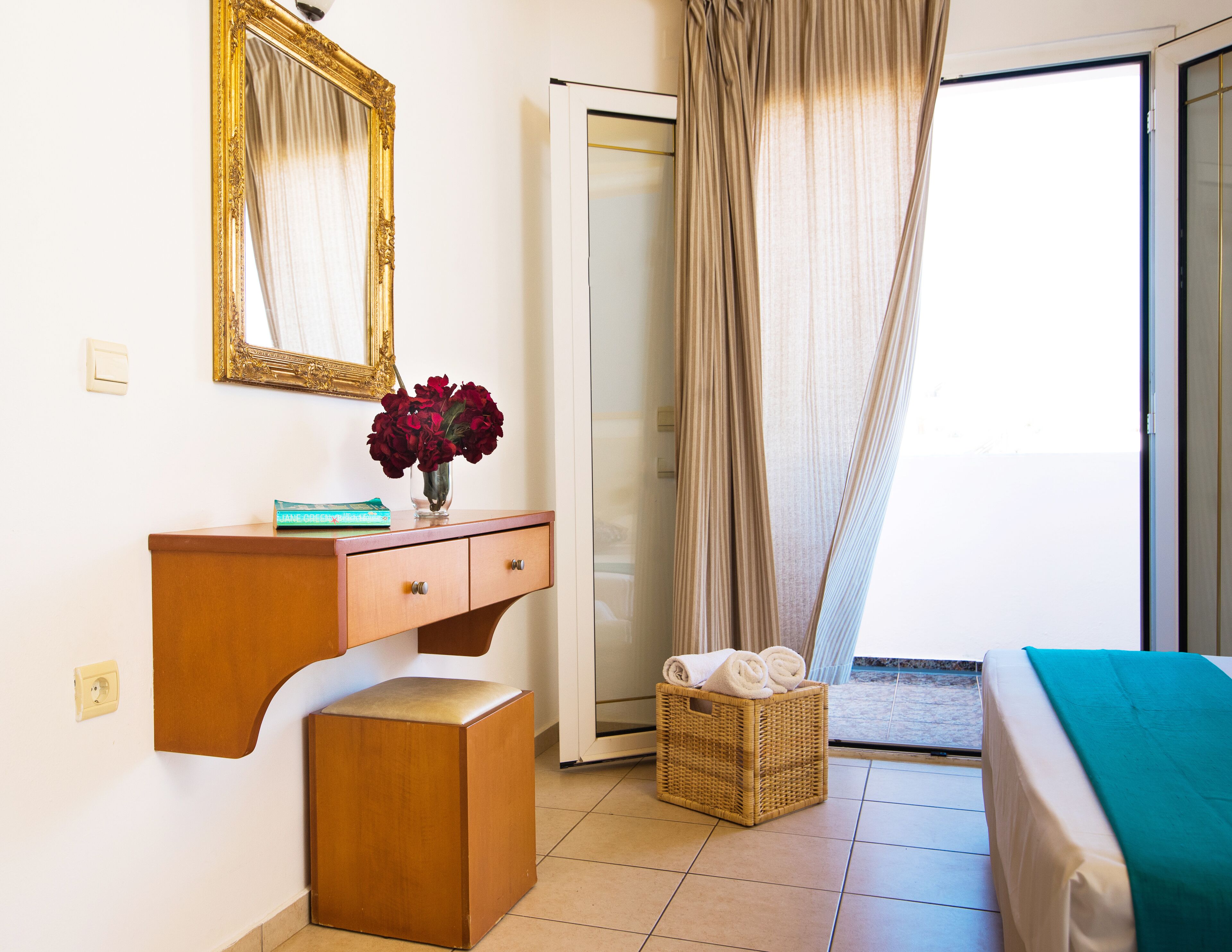 A hotel room with a wooden dresser, a mirror, a vase with flowers, a wicker basket with towels, and a bed with a green blanket.