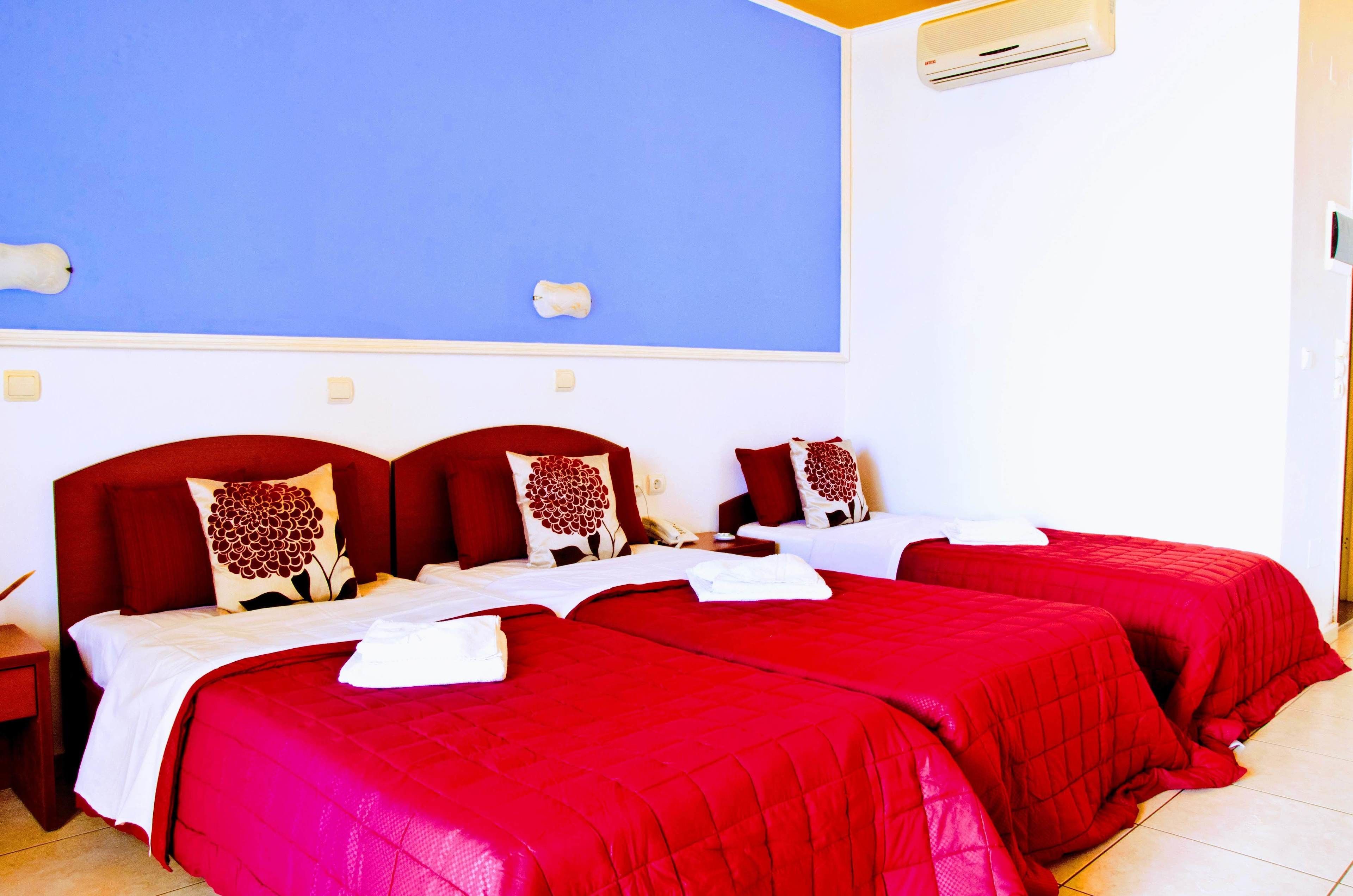 Habitación de hotel con dos camas, cada una con ropa de cama roja y almohadas decorativas, una mesita de noche blanca y una pared azul.