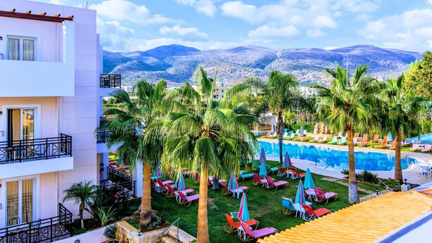 A pool area with palm trees, lounge chairs, and a mountain view. - Yiannis Manos (Hersonissos)