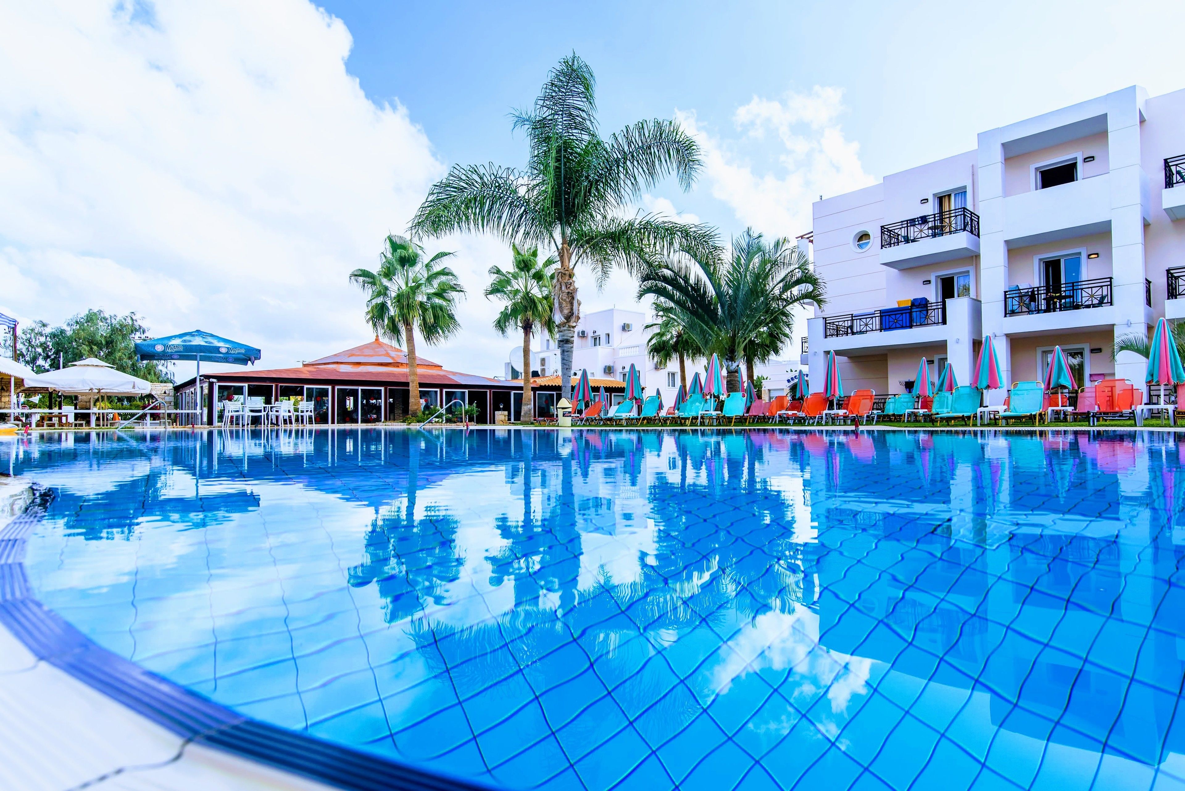 A swimming pool with clear blue water, surrounded by colorful lounge chairs and palm trees.