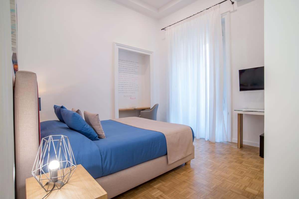 A bedroom with a bed, a desk, a chair, a TV, and a wall with a framed text.
