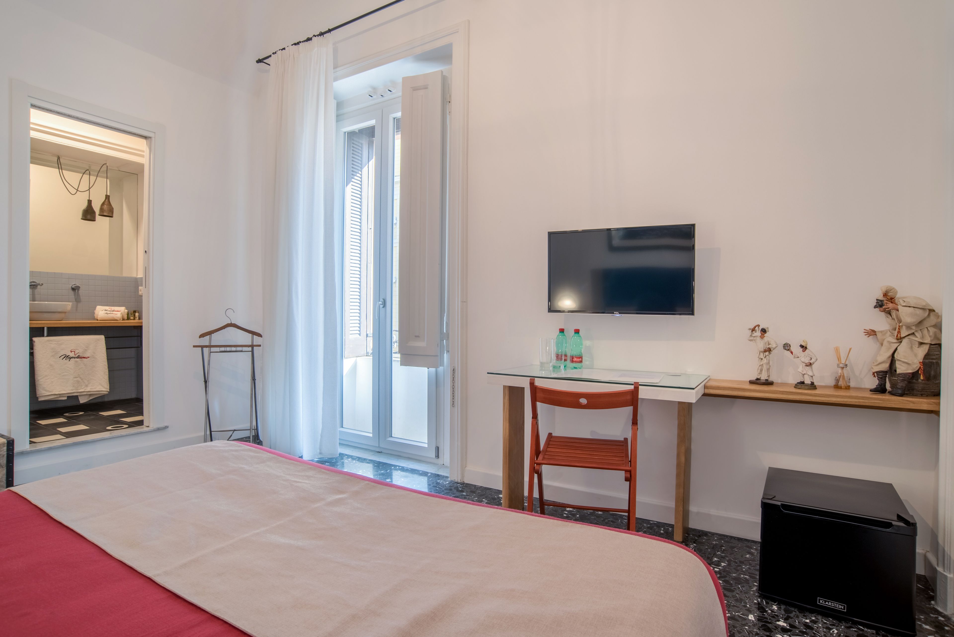 A hotel room with a bed, a desk with a chair, a television, a wardrobe, and a small shelf with figurines.