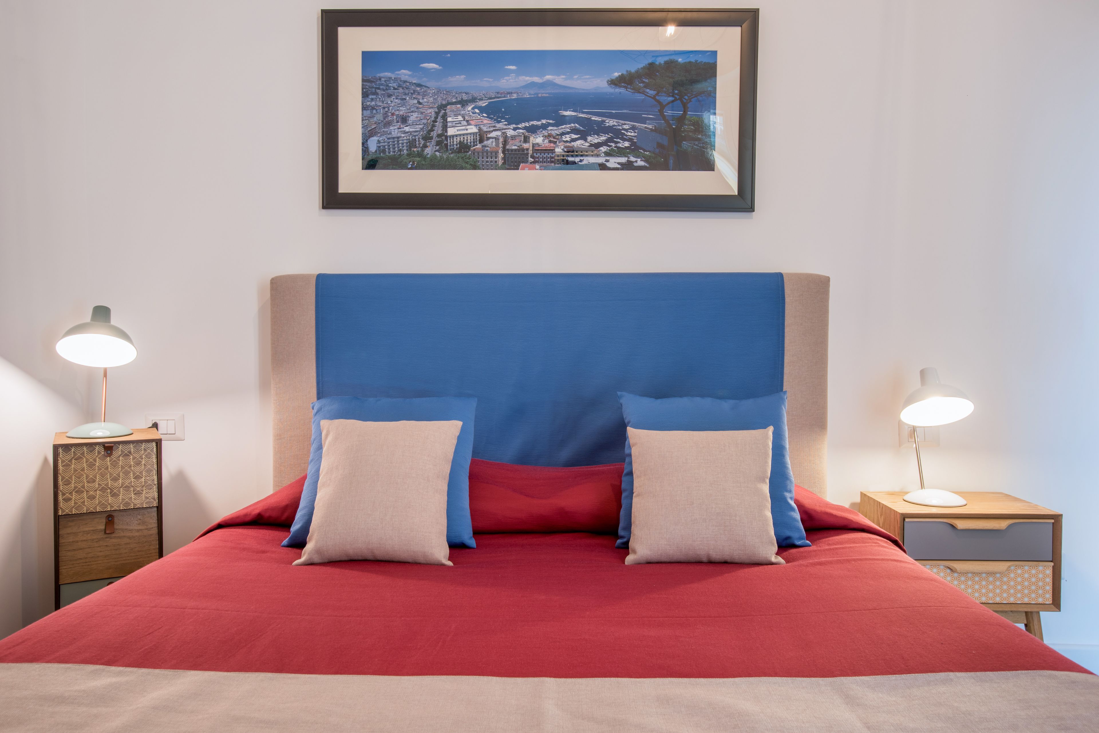 A bed with a red bedspread, two pillows, and a headboard with a cityscape view. There are two bedside tables with lamps.