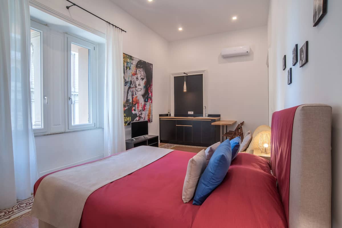 A hotel room with a bed, a desk, a chair, a window, and a painting on the wall.